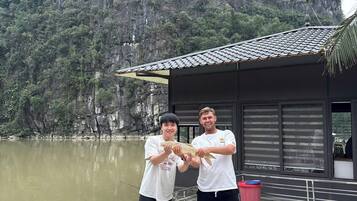 Dos personas sosteniendo un pez cerca de un edificio con techo de tejas y vista a un lago y montañas.
