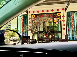Vue prise de l’intérieur d’une voiture, donnant sur un bâtiment décoré de lanternes rouges et d’éventails en papier, avec un arbre et un banc en bois au premier plan.