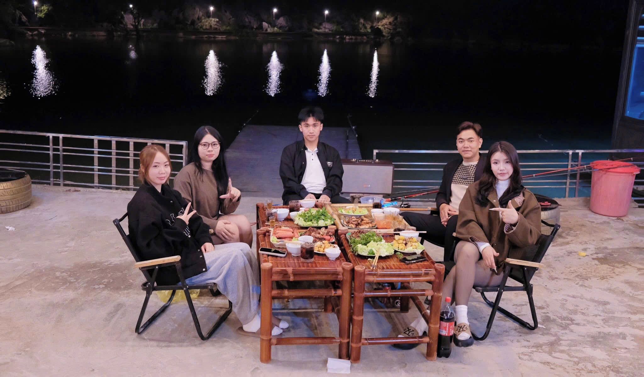 A group of people seated around a table with various dishes, outdoors at night with a water feature in the background.