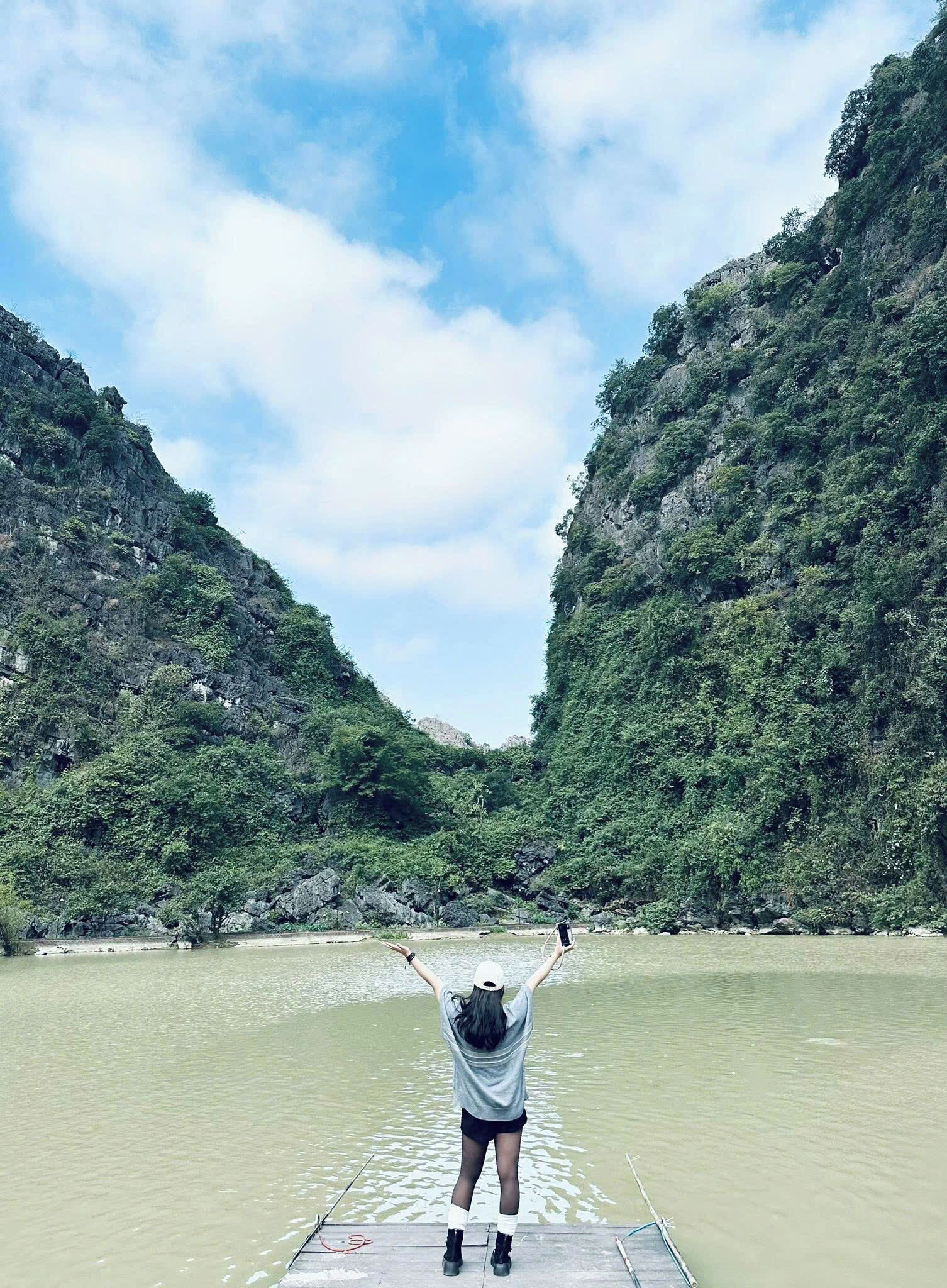 A person standing on a dock with arms raised, overlooking a river surrounded by lush green hills.