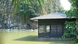 A cabin with a tiled roof, large windows, and a view of a calm river and forested cliffs. - Thien Truong An Eco Green Valley (Tay Hoa Lu)