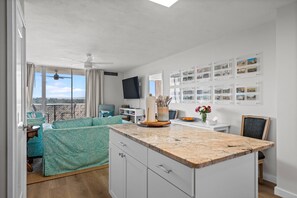 Ein modernes Wohnzimmer mit großem Fenster, Deckenventilator, Flachbildfernseher, Essbereich mit Tisch und Stühlen sowie einer Wand mit gerahmten Fotos.