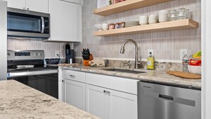 Una cocina moderna con gabinetes blancos, un microondas de acero inoxidable, un horno doble y un lavavajillas. Hay estantes de madera sobre el lavadero con platos y frascos.