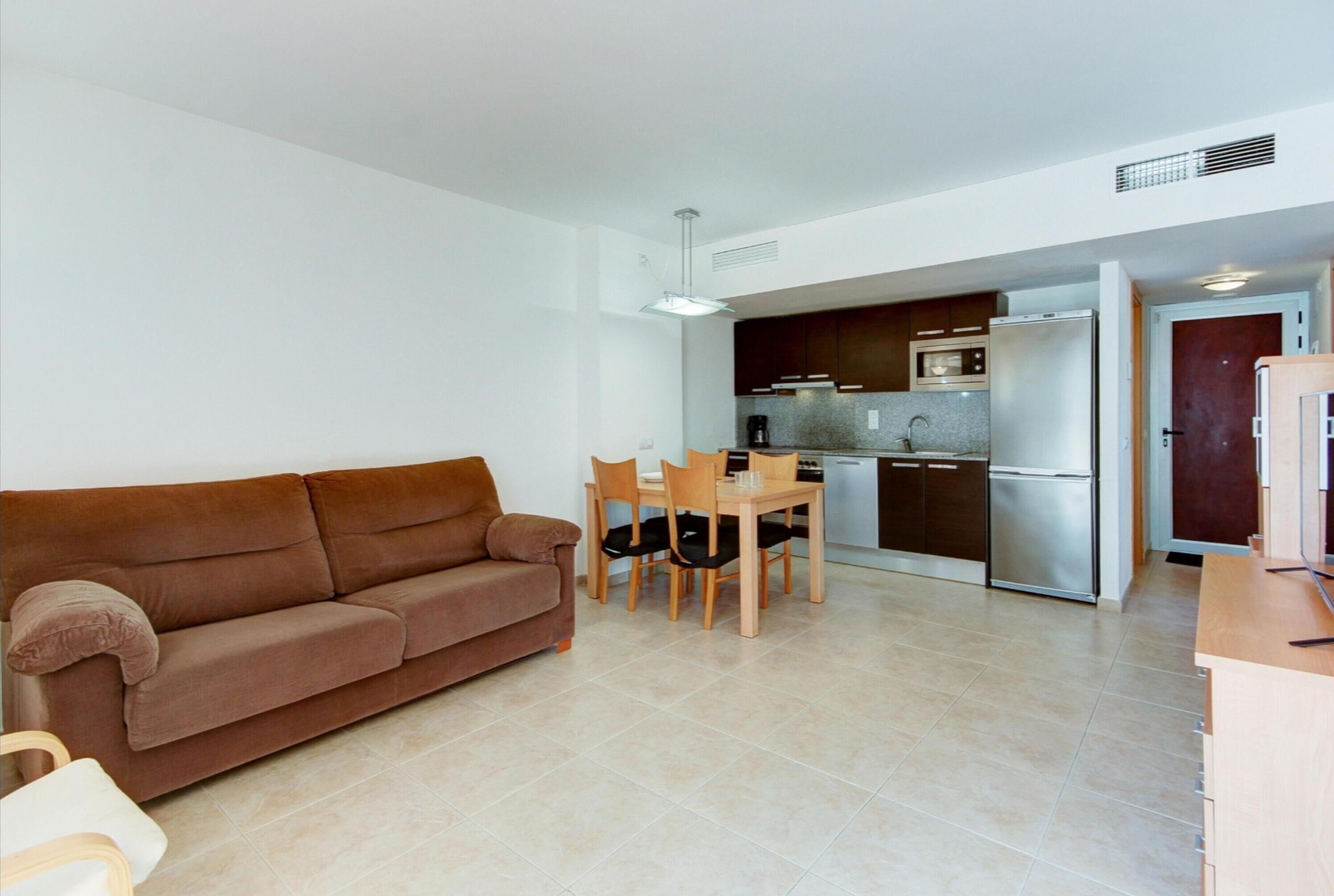 A modern living room with a brown sofa, a dining table with chairs, and a kitchen area with a refrigerator and microwave.