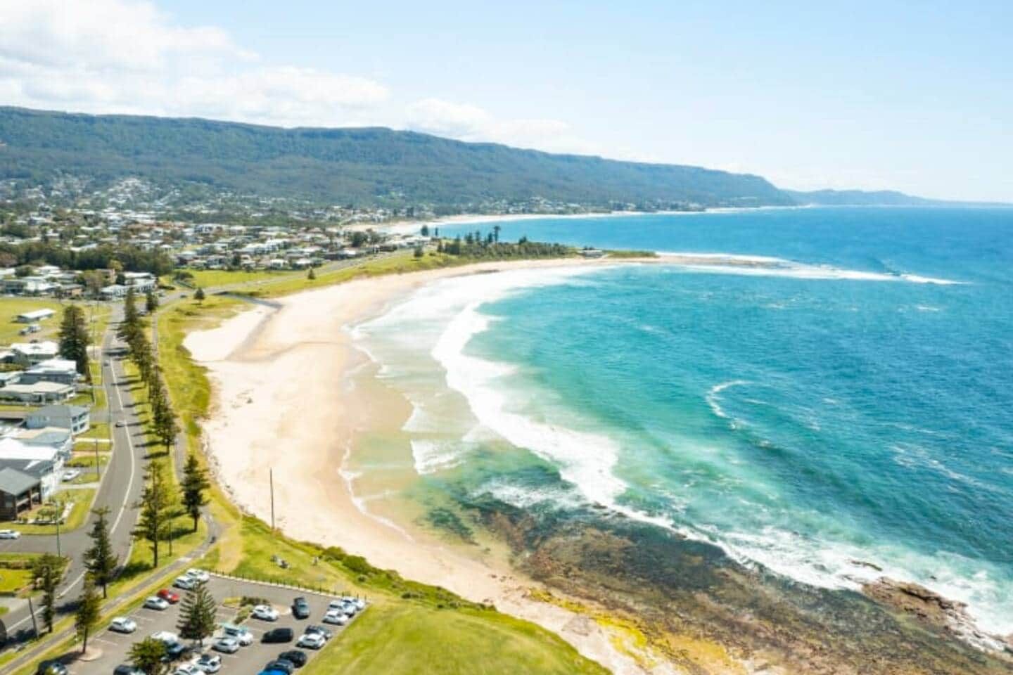 A coastal road with a beach and ocean on one side, and a residential area with houses and parked cars on the other.
