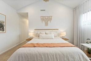 A bedroom with a large bed, two bedside tables with lamps, a macrame wall hanging, and a framed picture on the wall. - The White House - Beach Steps & Mountain Views (East Corrimal)