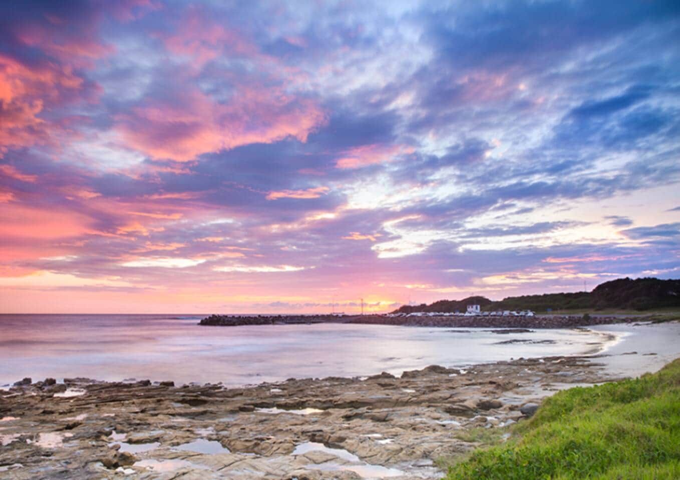 A coastal landscape with a rocky shoreline, a distant shoreline with buildings, and a sunset sky.