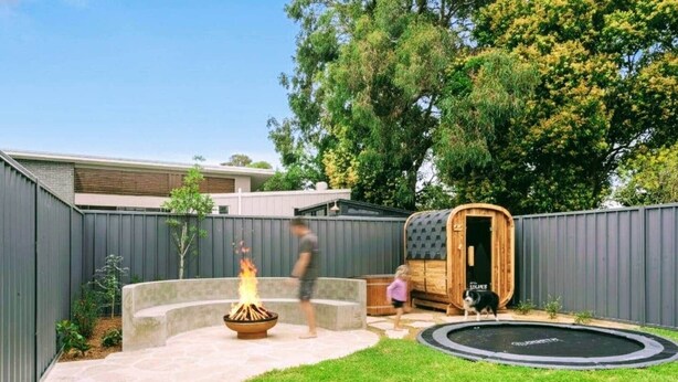 Un giardino con un braciere, una sauna in legno, un trampolino e un cane.