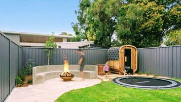 A backyard with a fire pit, a wooden sauna, a trampoline, and a dog.
