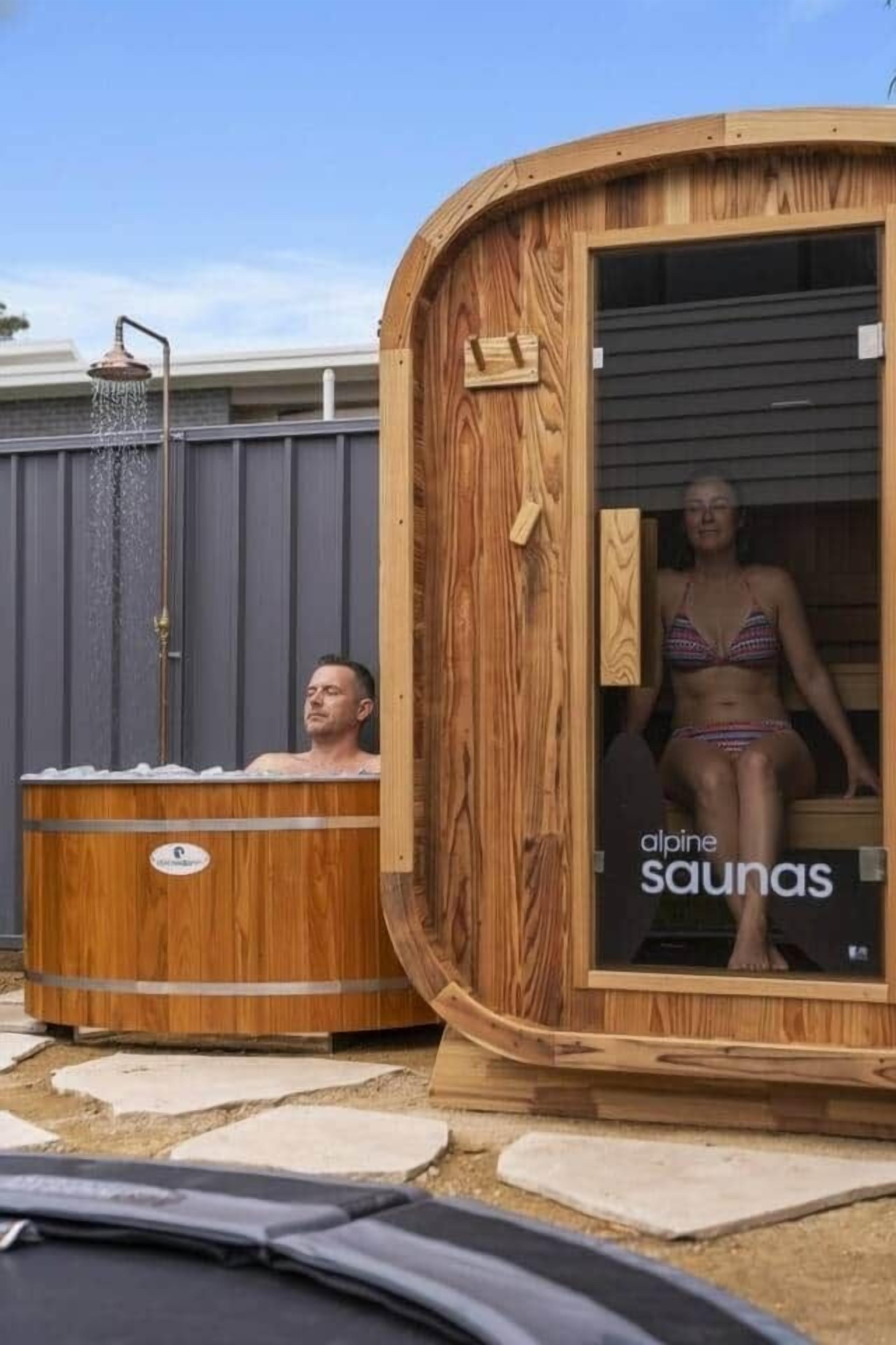 Twee houten sauna-cabines, met een douchekoof boven de één, en een bordje met de tekst 'Alpensauna's'.