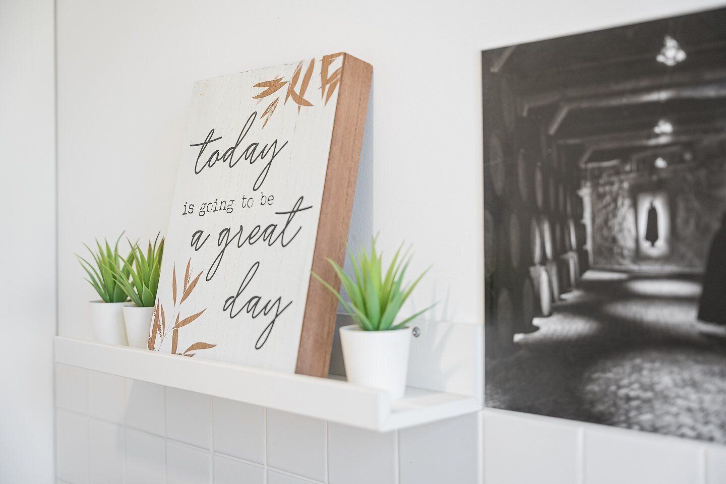 A wall-mounted sign with a positive message, flanked by two potted plants, and a black and white photograph to the right.