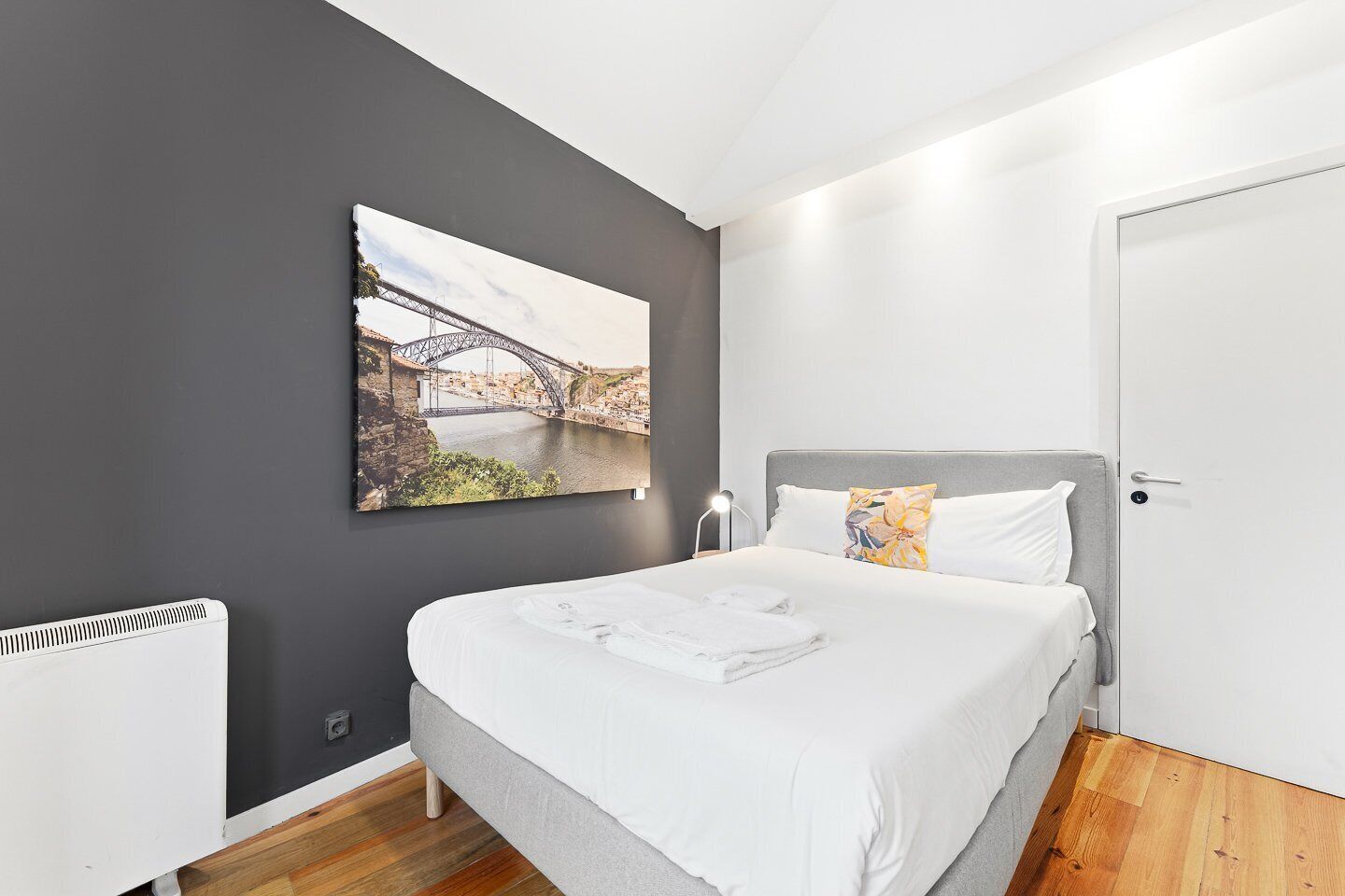 A bedroom with a bed, a nightstand, a lamp, a painting of a bridge, and a door.