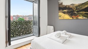A hotel room with a large window, a bed with white linens, a radiator, and a painting of a train on a bridge. - Guestready - Old City Design Apartments (Porto)