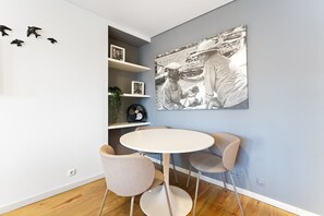 Un comedor con una mesa redonda y dos sillas, un estante en la pared con una planta, y una fotografía blanco y negro.