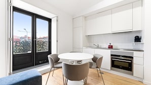 A modern kitchen with white cabinets, a round dining table with chairs, and a view through a large window. - Guestready - Old City Design Apartments (Porto)