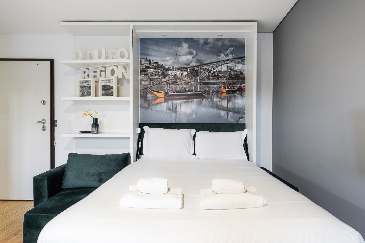 A modern hotel room with a large bed, a green sofa, a wall-mounted shelf with decorative items, and a cityscape painting.