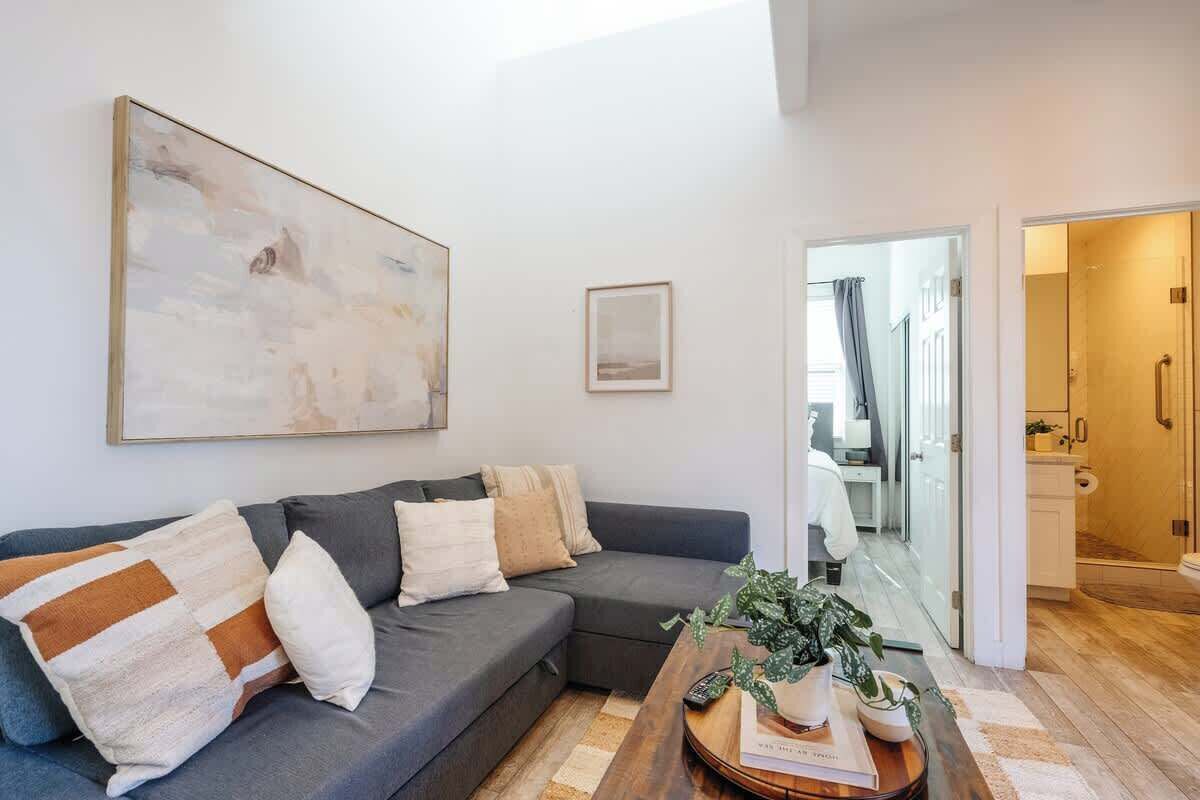 A modern living room with a grey sectional sofa, a coffee table with books and a plant, a large abstract painting, and a doorway leading to a bathroom.