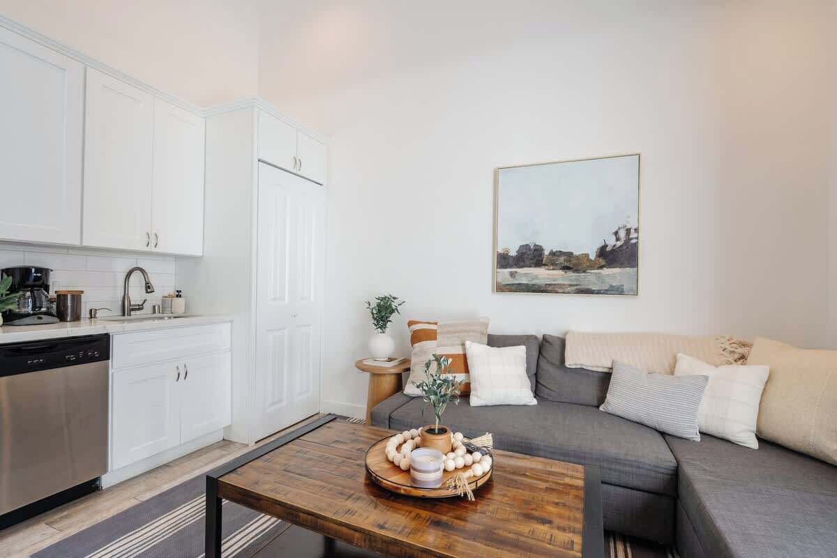 A modern kitchen with white cabinets, a dishwasher, and a coffee machine. There is a wooden coffee table with a tray of eggs and a vase. A gray sofa with cushions is in the living area, and a painting hangs on the wall.