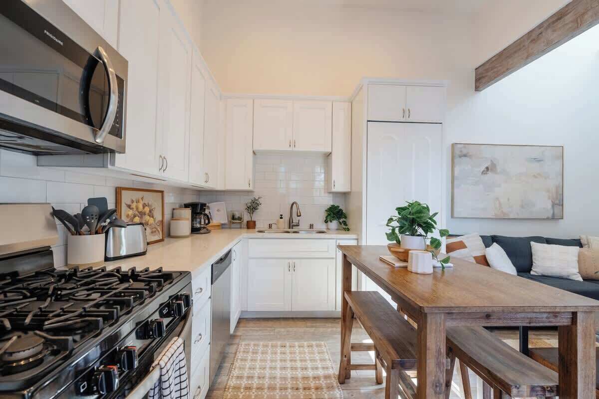 Une cuisine avec des meubles blancs, une cuisinière, un four à micro-ondes, un évier et un coin repas comprenant une table et un banc en bois.