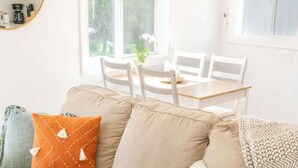 Un salon avec un canapé beige, une table basse ronde et une table à manger avec des chaises. Une fenêtre offre une vue sur la verdure à l’extérieur.