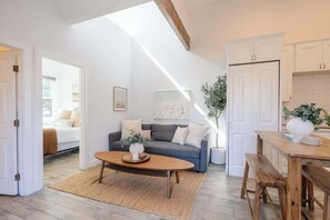 A modern living room with a sofa, coffee table, and a dining area with a bench. There is a large window providing natural light and a view of the outside.