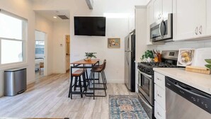 Een moderne keuken met witte kasten, roestvrijstalen apparatuur, een eethoek met een houten tafel en stoelen, en een groot raam dat natuurlijk licht binnenlaat.