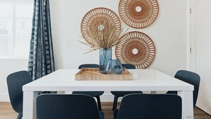 Un comedor con una mesa blanca, sillas oscuras, un florero azul con plantas secas y dos adornos de pared tejidos.