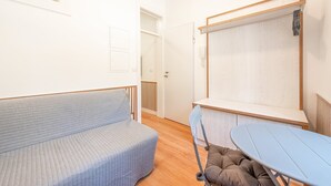 A room with a sofa, a table, a chair, a wooden floor, and a white wall with a door. - Guestready - Urban Garden Apartments (Lisbon)