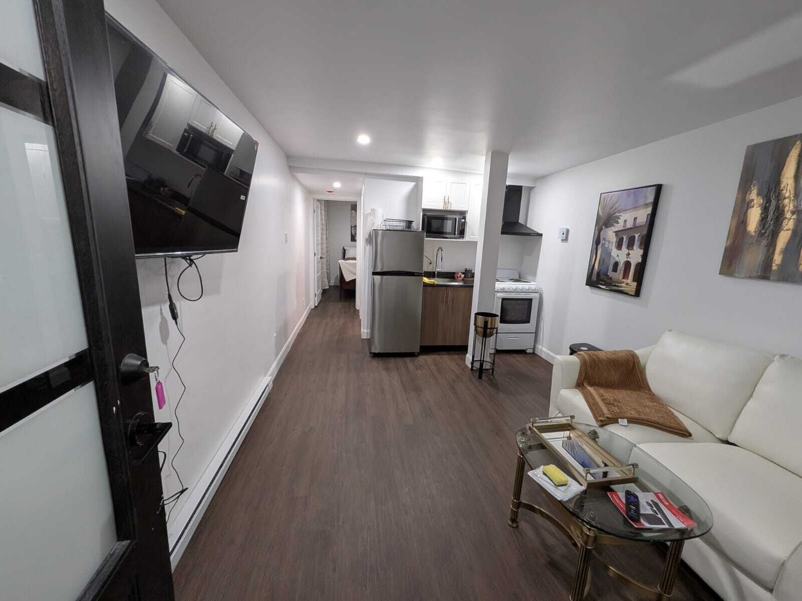 A modern living room with a white sofa, a glass coffee table, and a TV mounted on the wall. A kitchen with a refrigerator, microwave, and stove is visible in the background.