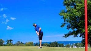 Una persona su un campo da golf con una bandiera rossa, una pallina da golf e un cielo blu limpido.