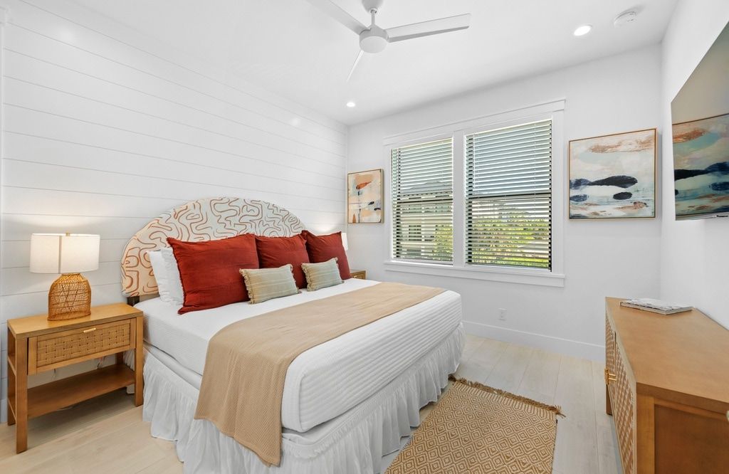 A bedroom with a bed, bedside tables, a ceiling fan, a window with blinds, and artwork on the walls.