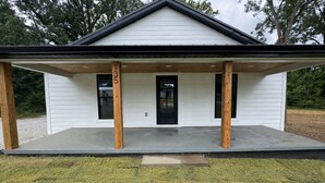 Un bâtiment blanc doté d’un porche couvert soutenu par des piliers en bois, d’une porte noire et d’une petite fenêtre.