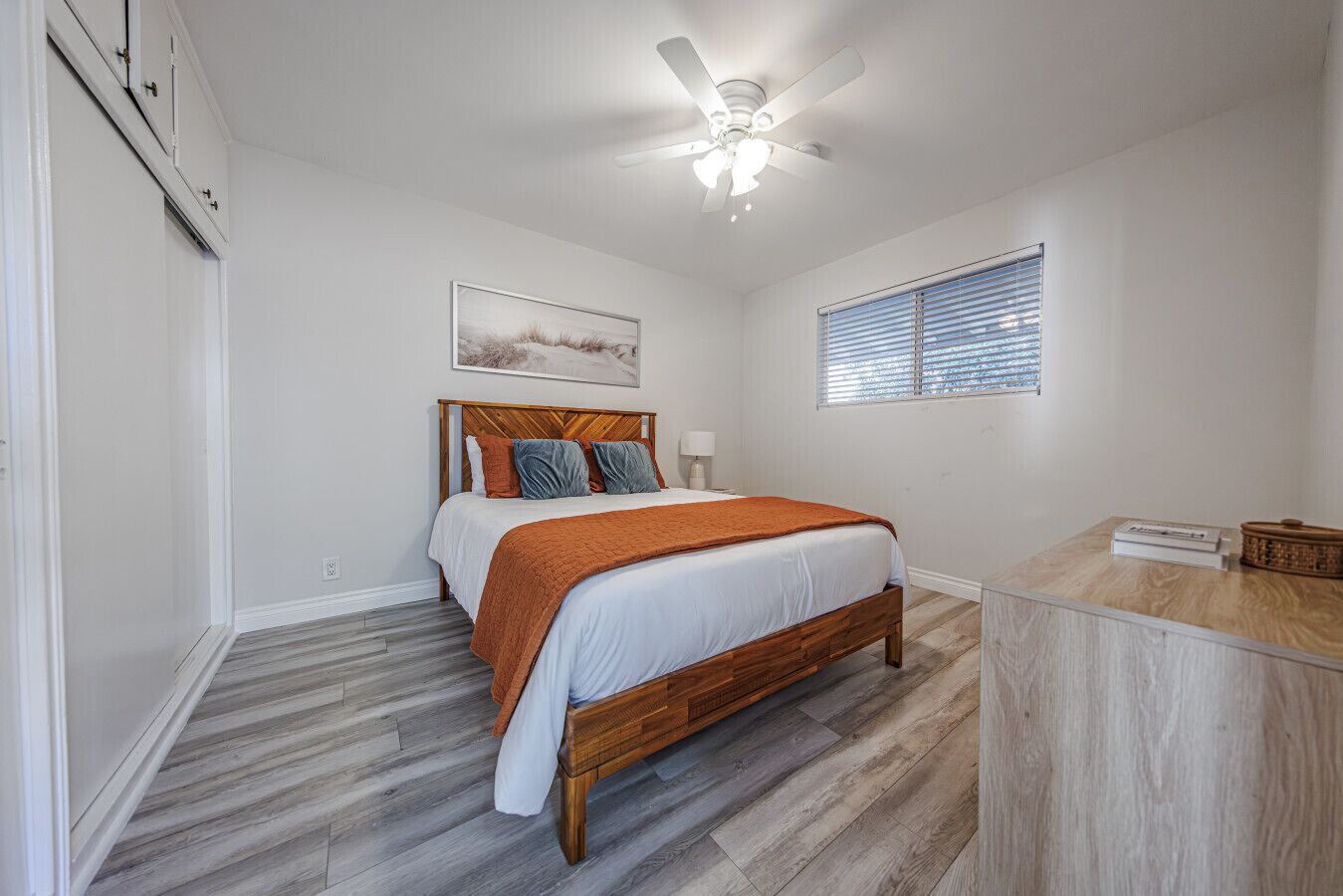 A bedroom with a wooden bed, a ceiling fan, a window with blinds, and a small table with a basket.
