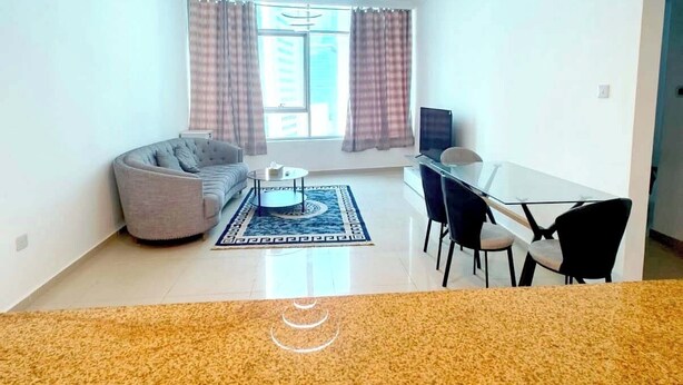 Uma sala de estar ampla, com sofá, mesa de jantar com cadeiras, mesa de centro e uma janela grande com cortinas.