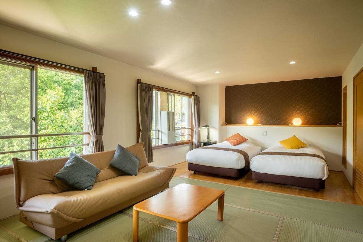 A hotel room with a sofa, two single beds, a wooden table, and large windows with curtains.