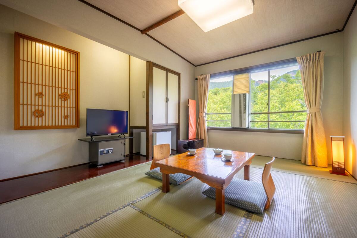 A traditional Japanese room with tatami flooring, a television, a wooden table, and a view of greenery through the window.
