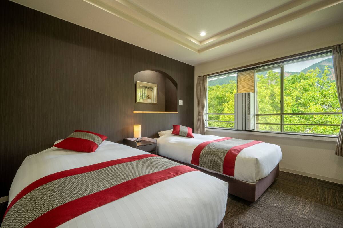A hotel room with two beds, a nightstand, a lamp, and a window with a view of greenery.
