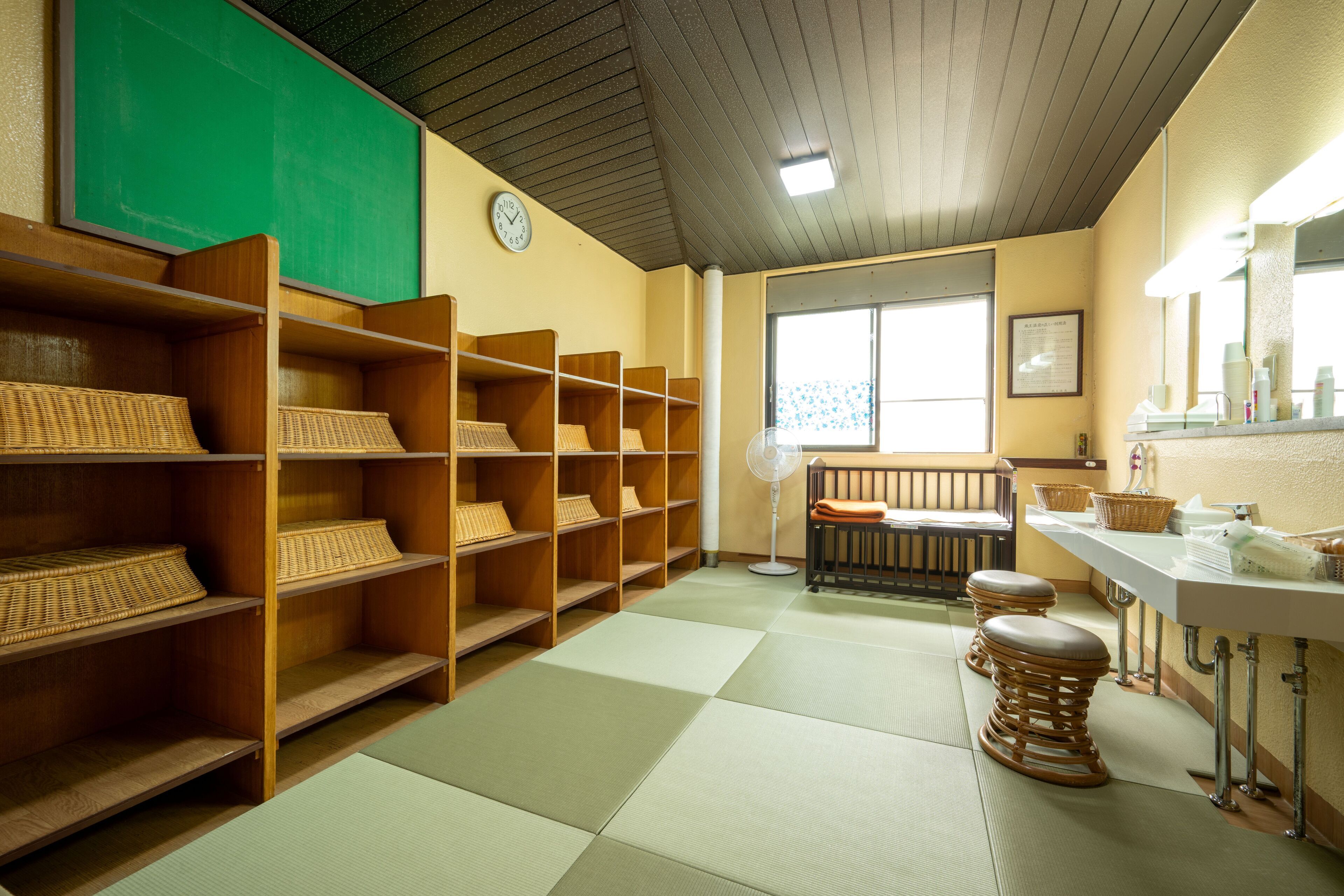 A library with wooden shelves, tatami mats, a fan, and a clock on the wall.