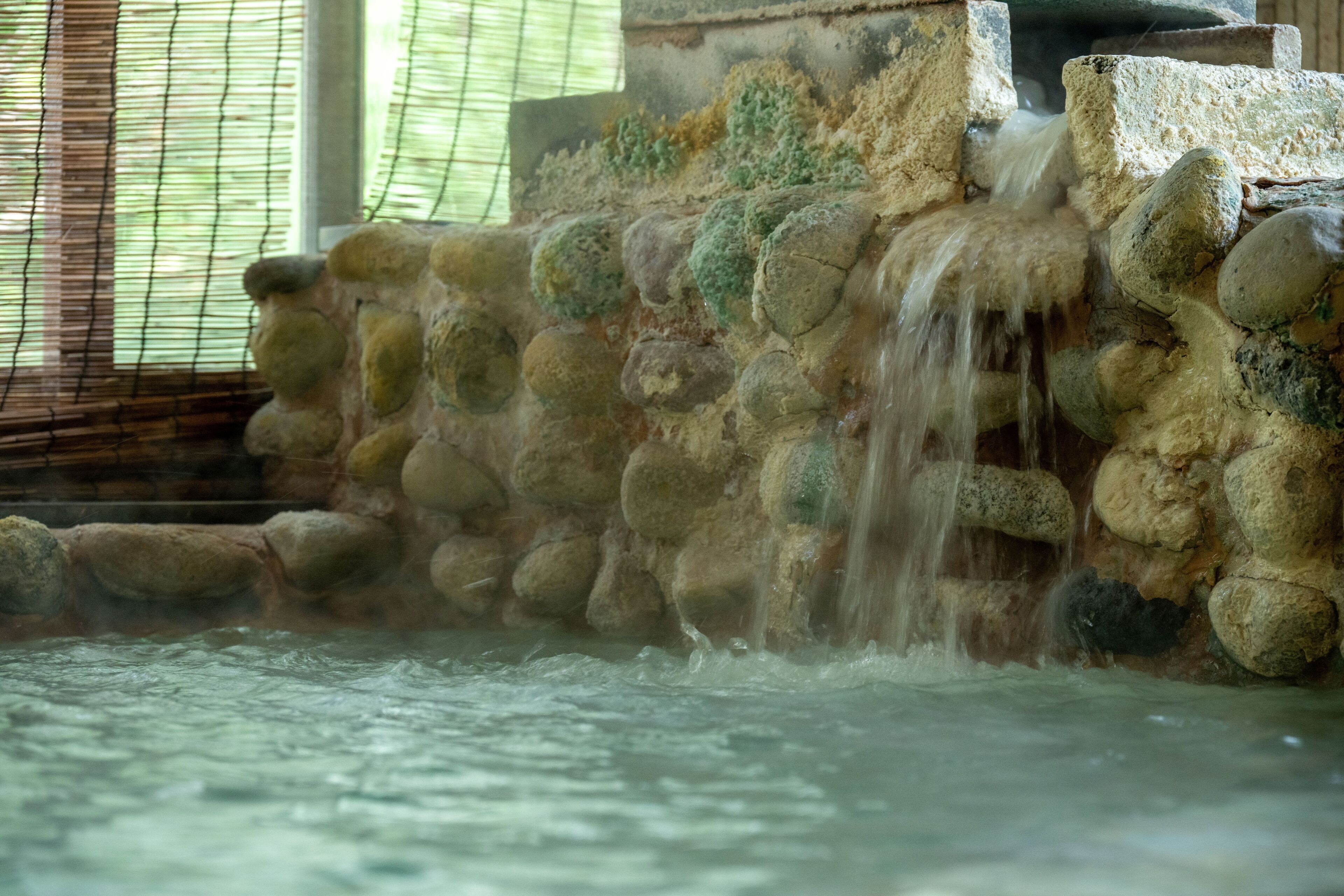 A stone wall with water cascading down, a window with blinds, and a stone structure in the background.