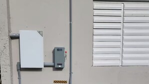 Ein Hauswirtschaftsraum mit zwei weißen Stromverteilern, einem Verteilerkasten und einem Fenster mit horizontalen Rollläden.