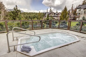 Un bain à remous avec une échelle en métal et une serviette posée sur le bord, entouré d’une rambarde en verre, offrant une vue sur une forêt et des bâtiments en arrière-plan.