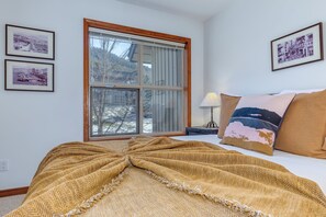 Une chambre avec une grande fenêtre, un lit recouvert d’une couverture à motifs et trois tableaux encadrés accrochés au mur.