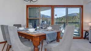 Ein Essbereich mit Holztisch, gedeckten Plätzen für vier Personen, grauen Polsterstühlen und Blick auf eine Berglandschaft durch große Fenster.