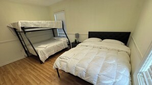 A room with a bunk bed and a single bed, wooden flooring, a nightstand with a lamp, and a window with blinds. - Modern 2BR Suite 202: Toledo & Sylvania Gateway (Toledo)