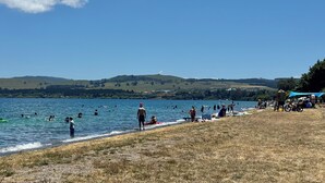 Une plage où l’on voit des gens se baigner et se promener le long du rivage, une zone herbeuse avec des tentes et, au loin, des collines.