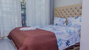 A bed with a tufted headboard, a red textured bedspread, and blue floral pillows. - golden leaf apartment (Kigali)
