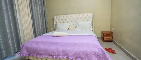 A neatly made bed with a purple bedspread and a white pillow, a wooden nightstand with a drawer, and a pair of red slippers on the floor.