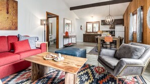 A spacious living room with a red sofa, a grey armchair, and a wooden coffee table. There is a kitchen in the background with wooden cabinets and a hanging light fixture.