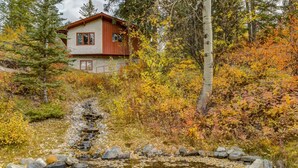 A rustic cabin with a stone foundation and wooden exterior, surrounded by autumn foliage and a small stream.