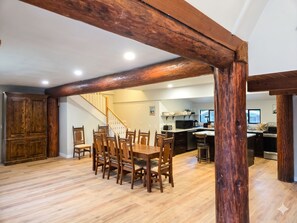 A spacious dining area with wooden chairs and a table, a kitchen in the background, and a staircase leading to another level.